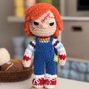 Könnte beinhalten: Handgemachte Häkelpuppe einer Figur mit orangefarbenem Haar, genähten Narben und schwarzen Augen. Die Puppe trägt eine blaue Latzhose, ein gestreiftes Hemd und rote Schuhe. Die Puppe wird auf einem Holzständer ausgestellt.