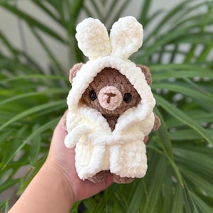 May include: A brown crocheted bear plush toy wearing a white hooded bathrobe with bunny ears. The bear has dark round eyes and a light brown stitched nose. The plush is made of soft, textured yarn and features a tie-front robe design.