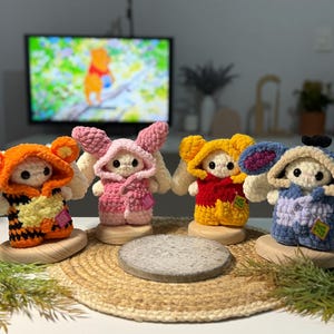 May include: Four handmade crochet bunny dolls, each wearing a different outfit. The bunnies are white with various colored outfits, including orange, pink, yellow, and blue. They are displayed on wooden bases and a woven mat.