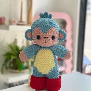 Puede incluir: Un peluche de mono tejido a ganchillo con cuerpo azul, cara y centros de orejas rosa claro, una mancha amarilla en el vientre y pies de color rojo brillante. El juguete tiene dos ojos de plástico negro, nariz y boca bordadas en marrón y un mechón.