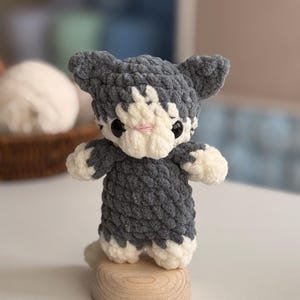 Puede incluir: Una figura de gato de ganchillo hecha a mano en gris y blanco, con ojos negros y nariz rosa. El peluche est&aacute; sobre una peque&ntilde;a base de madera. El gato tiene una textura esponjosa y es un objeto decorativo encantador.