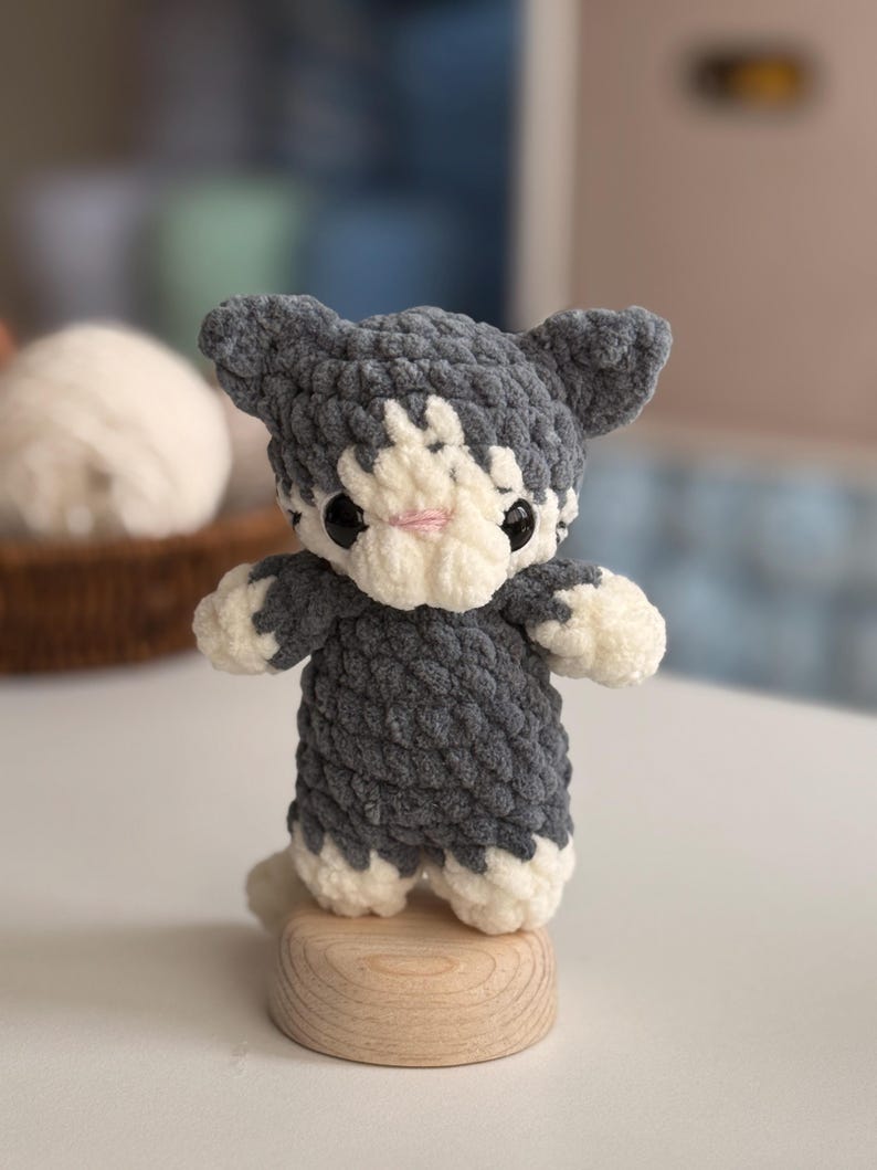 Puede incluir: Una figura de gato de ganchillo hecha a mano en gris y blanco, con ojos negros y nariz rosa. El peluche est&aacute; sobre una peque&ntilde;a base de madera. El gato tiene una textura esponjosa y es un objeto decorativo encantador.