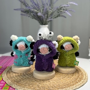 May include: Three handmade crochet cow figurines in teal, purple, and green hooded coats. Each cow has a white face, black ears, and a pink nose. They stand on wooden bases, with a woven mat in the foreground and a vase of lavender in the background.