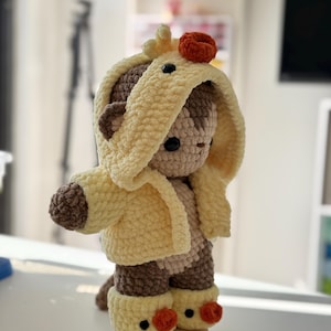 May include: A handmade crochet teddy bear wearing a yellow hooded jacket and matching boots. The bear has brown accents and a small orange detail on its head. The bear is standing on a wooden base.