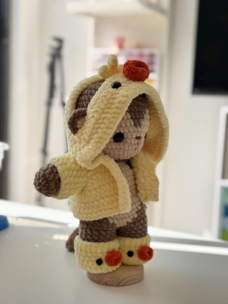 May include: A handmade crochet teddy bear wearing a yellow hooded jacket and matching boots. The bear has brown accents and a small orange detail on its head. The bear is standing on a wooden base.