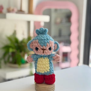 Op de afbeelding: Een gehaakt apenknuffeltje met een blauwe kop en lichaam, een lichtroze gezicht en handen, een lichtgele buik en felrode voeten. Het speelgoed heeft donkerbruin geborduurde ogen en een neus en staat rechtop op een kleine houten voet.