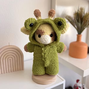 Puede incluir: Una muñeca jirafa de ganchillo hecha a mano con un disfraz de rana verde. La jirafa tiene un cuerpo amarillo y marrón con un hocico blanco y ojos negros. El disfraz de rana incluye una capucha con ojos grandes y un mono con botones.