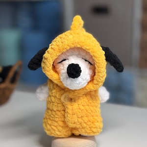 Puede incluir: Muñeco Snoopy de ganchillo hecho a mano con un chaleco con capucha amarillo. El muñeco tiene orejas negras, una nariz negra y los ojos cerrados. El chaleco tiene un cierre de un solo botón. El muñeco se exhibe sobre un pequeño soporte de madera.