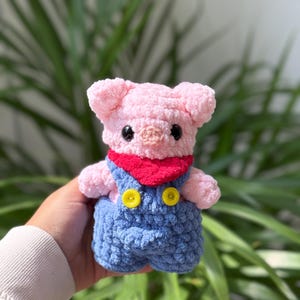 以下が含まれることがあります： 柔らかいピンクの毛糸で編まれた手作りの豚のぬいぐるみ。黒いプラスチックの目、小さな鼻、赤いネッカチーフ、そして服の前のストラップに2つの明るい黄色のボタンが付いた青いオーバーオールが特徴です。