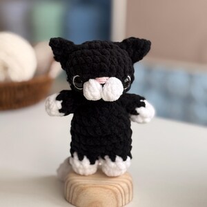 Puede incluir: Figura de gato de ganchillo en blanco y negro. El gato tiene patas blancas, hocico blanco y nariz rosa. Est&aacute; sobre una peque&ntilde;a base redonda de madera. Los ojos del gato son negros.