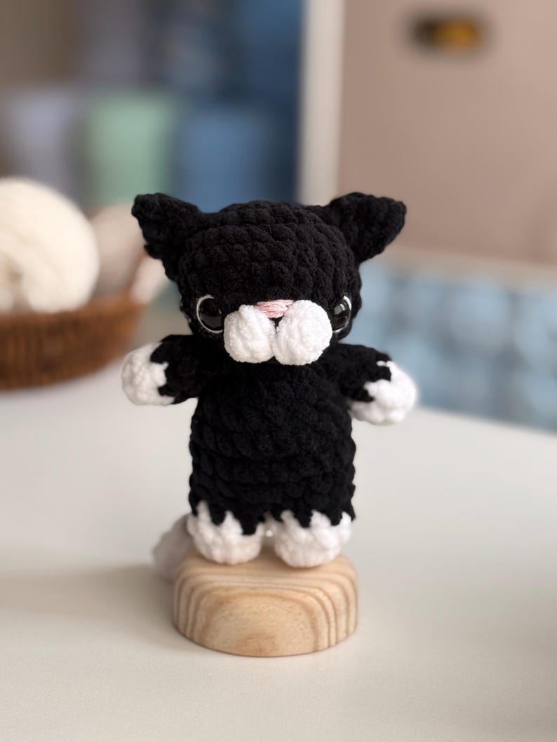 Puede incluir: Figura de gato de ganchillo en blanco y negro. El gato tiene patas blancas, hocico blanco y nariz rosa. Est&aacute; sobre una peque&ntilde;a base redonda de madera. Los ojos del gato son negros.
