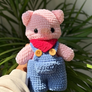 Puede incluir: Un cerdo de peluche tejido a mano con hilo rosa claro suave, ojos de seguridad negros y un hocico pequeño. El cerdo lleva un pañuelo rojo brillante y un peto azul con dos botones amarillos en los tirantes y un detalle de bolsillo frontal.