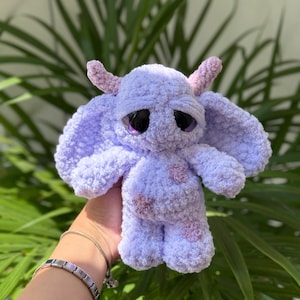 Puede incluir: Una criatura de peluche tejida a ganchillo en color violeta claro con ojos grandes, oscuros y brillantes, dos pequeños cuernos texturizados y orejas caídas. El cuerpo es de hilo suave y esponjoso con parches en forma de flor violeta oscuro.