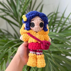 May include: A handmade crochet doll featuring dark blue yarn hair with a small yellow flower accessory, black button eyes, and a peach-colored face. The doll wears a yellow hooded jacket, a red skirt, striped leggings, and yellow boots with a red crossbody bag.
