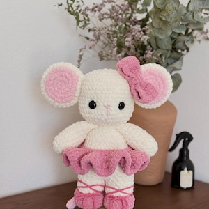May include: A handmade crochet mouse wearing a pink tutu and ballet slippers. The mouse has large, round ears with pink centers and a matching bow. The toy is white with black eyes and is displayed on a wooden base.