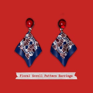Puede incluir: Un par de pendientes con una gema roja en la parte superior y un patrón de desplazamiento floral azul, blanco y marrón. Los pendientes tienen forma cuadrada y cuelgan de un gancho plateado. El texto "Floral Scroll Pattern Earrings" está en la parte inferior.