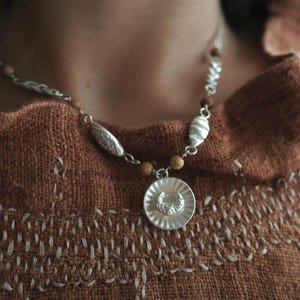 Op de afbeelding: Een zilveren ketting met een ronde hanger voorzien van een gestructureerd zonnestraalpatroon en een kleine halve maan in het midden. De ketting bestaat uit zilveren schakels afgewisseld met kleine, ronde, beige kralen en langwerpige zilveren decoratieve segmenten.