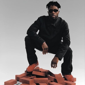 May include: A person wearing black clothing and sunglasses squats on a pile of red bricks. The person is wearing red sneakers. The background is a solid light gray.