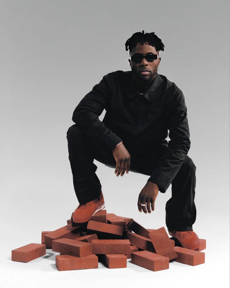 May include: A person wearing black clothing and sunglasses squats on a pile of red bricks. The person is wearing red sneakers. The background is a solid light gray.