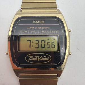 May include: A gold-tone Casio digital watch with a metal link band. The black watch face displays the time 7:30:56, the text CASIO, ALARM CHRONOGRAPH, ALARM, DUAL, TIMER, CHRONO, and the True Value logo. The watch case has a rectangular digital display.