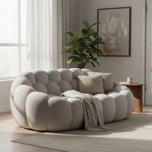 May include: A light gray, bubble-shaped sofa with two throw pillows and a blanket. The sofa is in a living room setting with a plant, artwork, and a small wooden side table. The room has a neutral color palette.