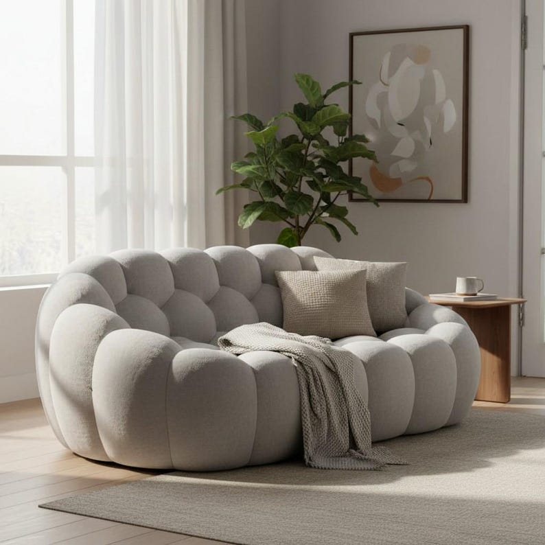 May include: A light gray, bubble-shaped sofa with two throw pillows and a blanket. The sofa is in a living room setting with a plant, artwork, and a small wooden side table. The room has a neutral color palette.
