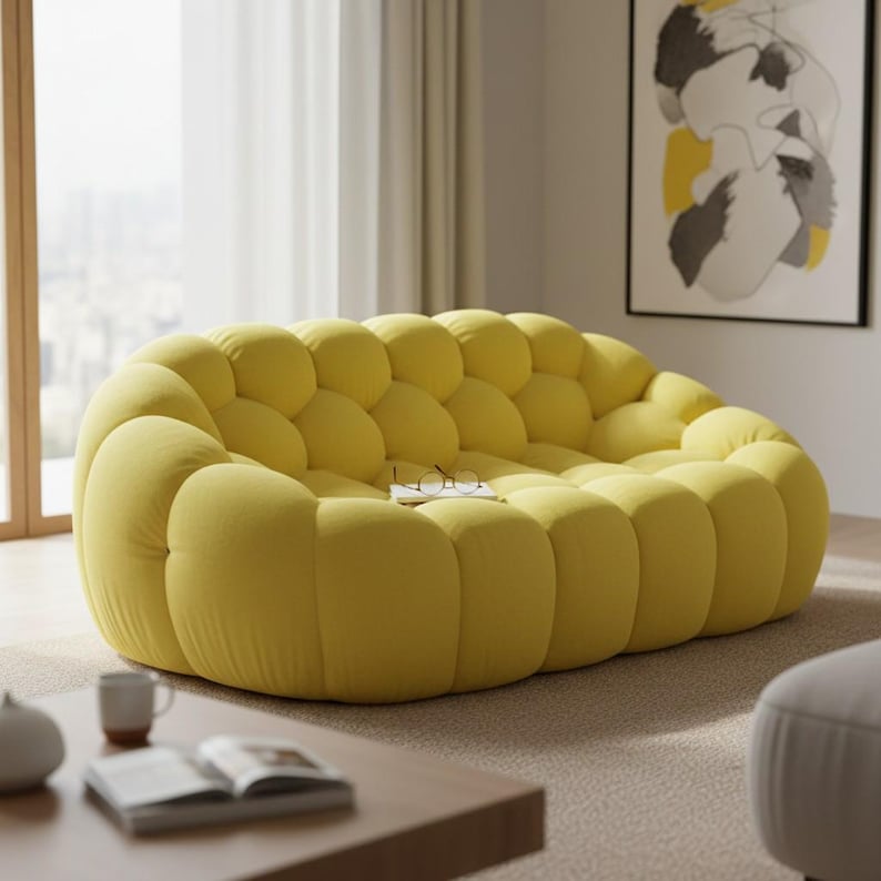 May include: A large, yellow, bubble-shaped sofa. The sofa has a rounded design with multiple individual cushions, creating a textured appearance. A book and glasses rest on the sofa. The sofa is in a living room setting with a neutral color scheme.