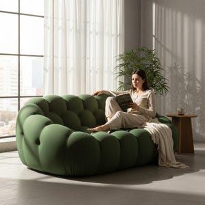 May include: An olive green, bubble-shaped sofa. The sofa has a rounded design with multiple individual cushion-like sections. A person is seated on the sofa, reading a book. A neutral-colored blanket is draped over the sofa. A small wooden side table and a plant are in the background.