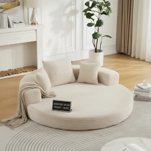 May include: A cream-colored, round sofa with a corduroy texture. It includes several matching pillows and a throw blanket. The sofa's diameter is 70 inches and weighs 330 pounds. The sofa is in a living room setting.