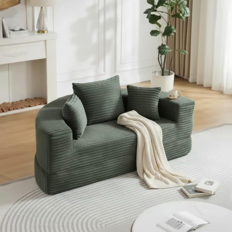 May include: A green corduroy sofa with three matching pillows and a cream-colored throw blanket. The sofa has a rounded shape and is placed on a patterned rug. A small side table and a potted plant are also visible.
