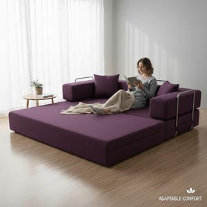May include: A modular, plum-colored sofa with a metal frame, shown in a living room setting. The sofa is in a flat, bed-like configuration, with a person reading a book. The sofa has several matching throw pillows and a blanket.