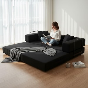 May include: A black convertible sofa bed with a gray throw blanket. The sofa has black cushions and metal armrests. A person is sitting on the sofa reading a book. A coffee mug and a stack of books are on the floor.