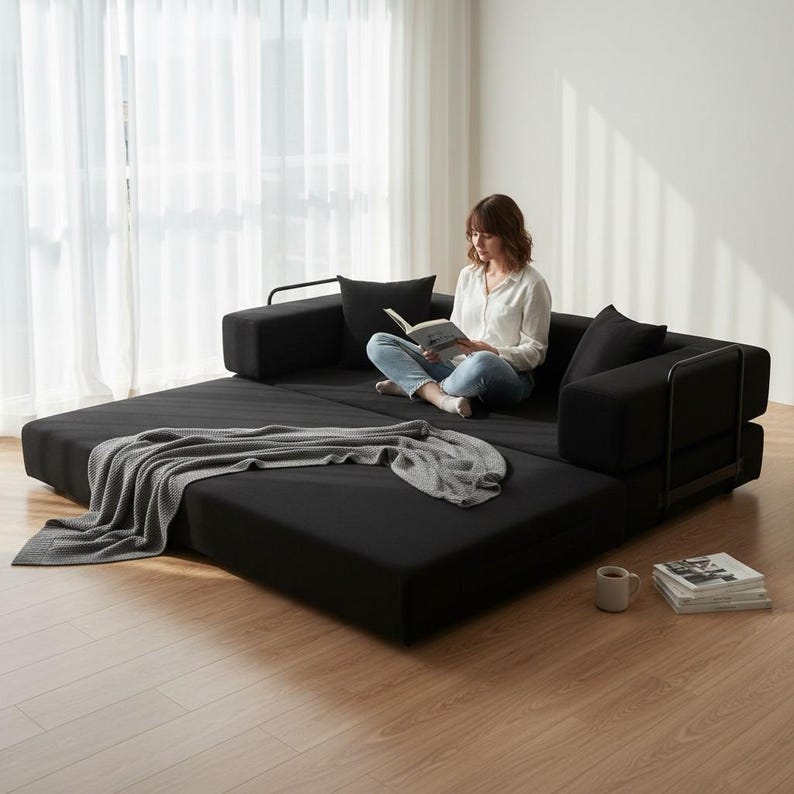 May include: A black convertible sofa bed with a gray throw blanket. The sofa has black cushions and metal armrests. A person is sitting on the sofa reading a book. A coffee mug and a stack of books are on the floor.