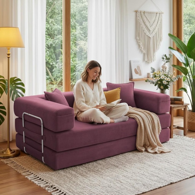 May include: A modular, burgundy corduroy sofa with a person sitting on it reading a book. The sofa has a rectangular shape with armrests and a metal frame. A beige throw blanket and pillows add comfort. The room has a neutral color palette.