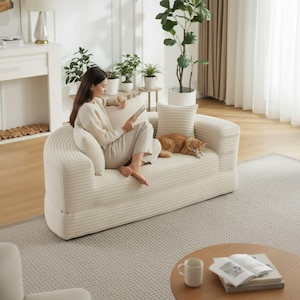 May include: A cream-colored corduroy sofa with rounded arms and back. A woman sits on the sofa, holding a tablet. A ginger cat rests on the sofa. The room has a neutral color palette with plants and a wooden coffee table.