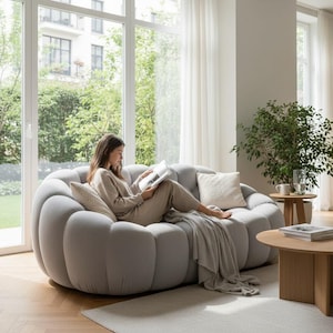 May include: A light gray, modern sofa with a unique, rounded design. The sofa has a soft, textured appearance and is paired with decorative pillows and a throw blanket. A person is seated on the sofa, reading a book in a bright, airy living room.