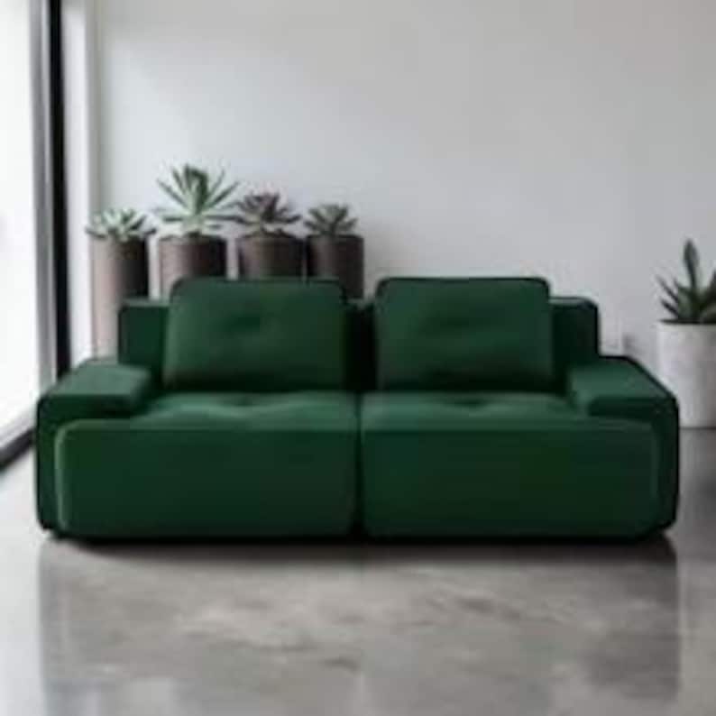 Puede incluir: Un sof&aacute; moderno de color verde esmeralda con un dise&ntilde;o minimalista. El sof&aacute; tiene dos cojines grandes para el asiento y dos cojines para el respaldo, con reposabrazos a ambos lados. Est&aacute; colocado frente a una pared blanca y un suelo de hormig&oacute;n.