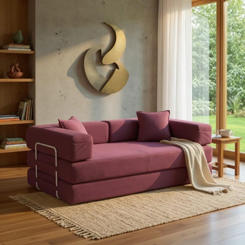 May include: A modern, modular sofa in a deep rose color with white metal accents. The sofa has a layered design and comes with matching throw pillows. A beige throw blanket rests on the sofa. The room features a large window and a decorative wall sculpture.