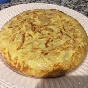 Könnte beinhalten: Eine goldbraune spanische Tortilla, ein traditionelles Gericht aus Eiern, Kartoffeln und Zwiebeln, wird auf einem weißen Teller mit dekorativem Rand präsentiert.