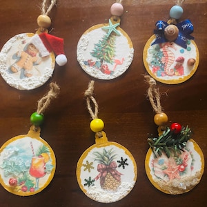 Puede incluir: Seis adornos navideños redondos con varios diseños. Uno presenta un hombre de jengibre con un gorro de Papá Noel, otro un árbol de Navidad y un flamenco, y un tercero un caballito de mar. Otros adornos tienen un cóctel, una piña y conchas.
