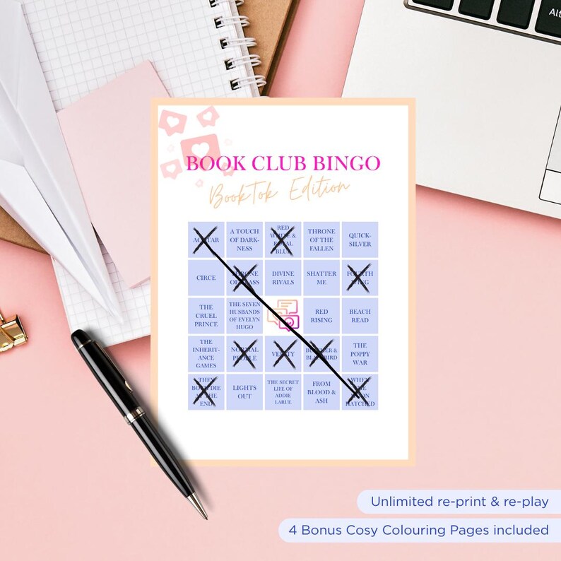 May include: A Book Club Bingo card with the text "Book Club Bingo BookTok Edition" in pink. The card has squares with book titles and crossed-out spaces. A black pen lies on the pink surface next to the card. The bottom of the card reads "Unlimited re-print & re-play".