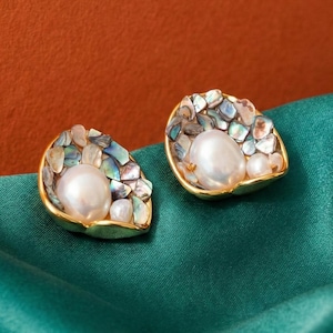 Può includere: Un paio di orecchini a bottone dorati. Ogni orecchino presenta una grande perla bianca rotonda circondata da frammenti di conchiglia iridescenti multicolori. Gli orecchini poggiano su un tessuto di raso verde acqua su uno sfondo arancione bruciato.