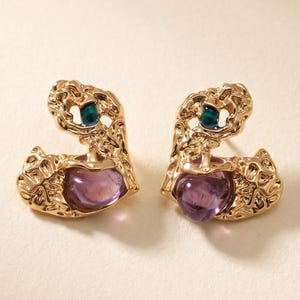 May include: A pair of gold-toned earrings with an irregular, textured metal frame. Each earring features a smooth, translucent purple stone at the base and a small, deep teal circular accent embedded within the upper section of the metal structure.