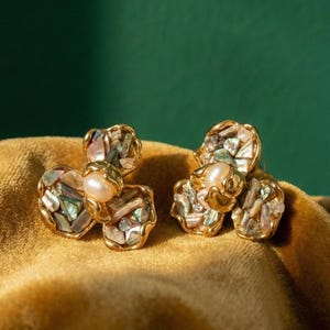 May include: A pair of gold-toned stud earrings shaped like three-petaled flowers. Each petal is inlaid with iridescent crushed shell fragments in shades of pink, green, and white, with a single round, cream-colored pearl set in the center.