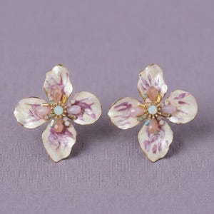 May include: A pair of floral stud earrings. Each earring features four white petals with purple veining. The center of each flower is adorned with pink, blue, and gold-colored gemstones. The earrings are set against a lavender background.