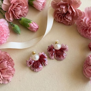 May include: A pair of flower-shaped earrings with a pearl center and gold accents, surrounded by pink carnations and buds. A cream-colored ribbon is also present, all on a light beige surface.