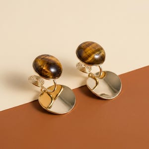 May include: A pair of clip-on earrings featuring polished, oval-shaped tiger's eye gemstones in shades of brown and gold. Each earring is attached to a gold-toned metal coil mechanism connected to a smooth, circular gold-toned metal disc base.