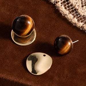 May include: A pair of stud earrings featuring polished, spherical tiger's eye gemstones in brown and gold tones. Each earring includes a detachable, smooth, circular gold-toned metal disc component that sits behind the earlobe for a layered look.