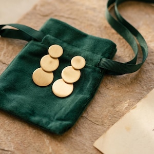 May include: A pair of gold-toned earrings featuring three vertically aligned circular discs of varying sizes with a brushed matte finish, resting on a small, dark green velvet drawstring pouch with a matching green ribbon tie.