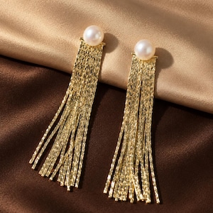 May include: A pair of gold-toned drop earrings featuring a single white pearl at the top of each piece. Attached to the base of each pearl is a series of thin, textured gold-coloured metal chains that hang vertically to create a fringe effect.